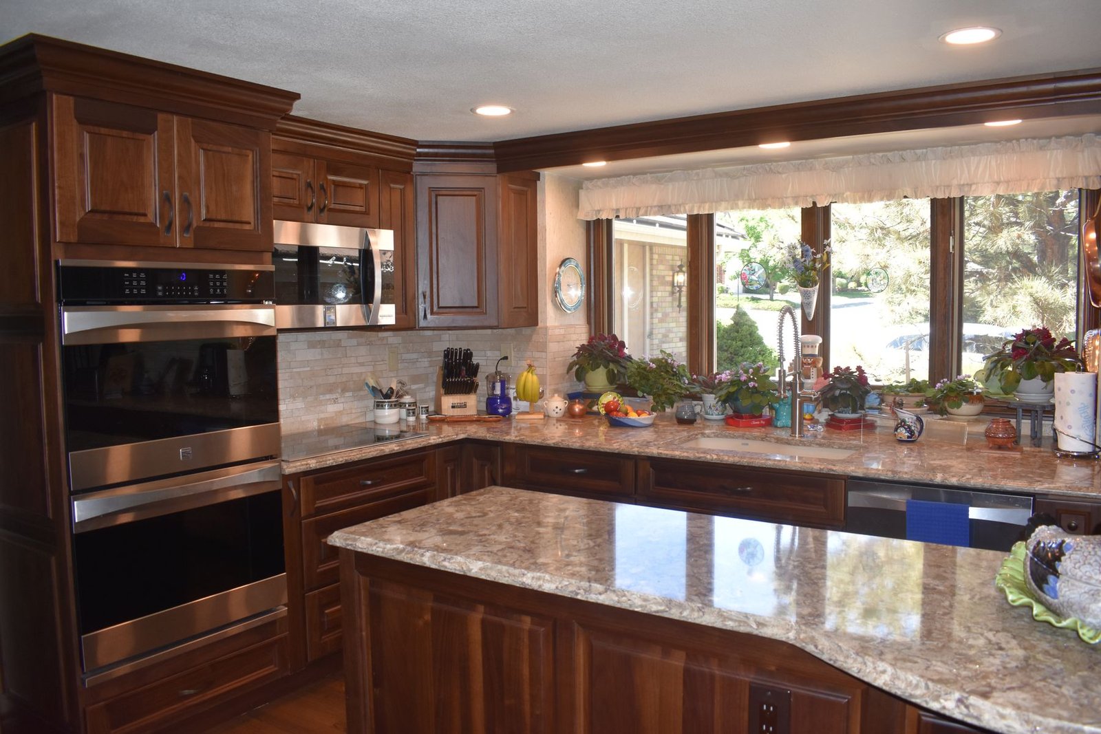 A finished kitchen with cherry raised panel cabinets, granite counters, stainless double wall ovens, a curved granite island and undercabinet lighting along a run of windows