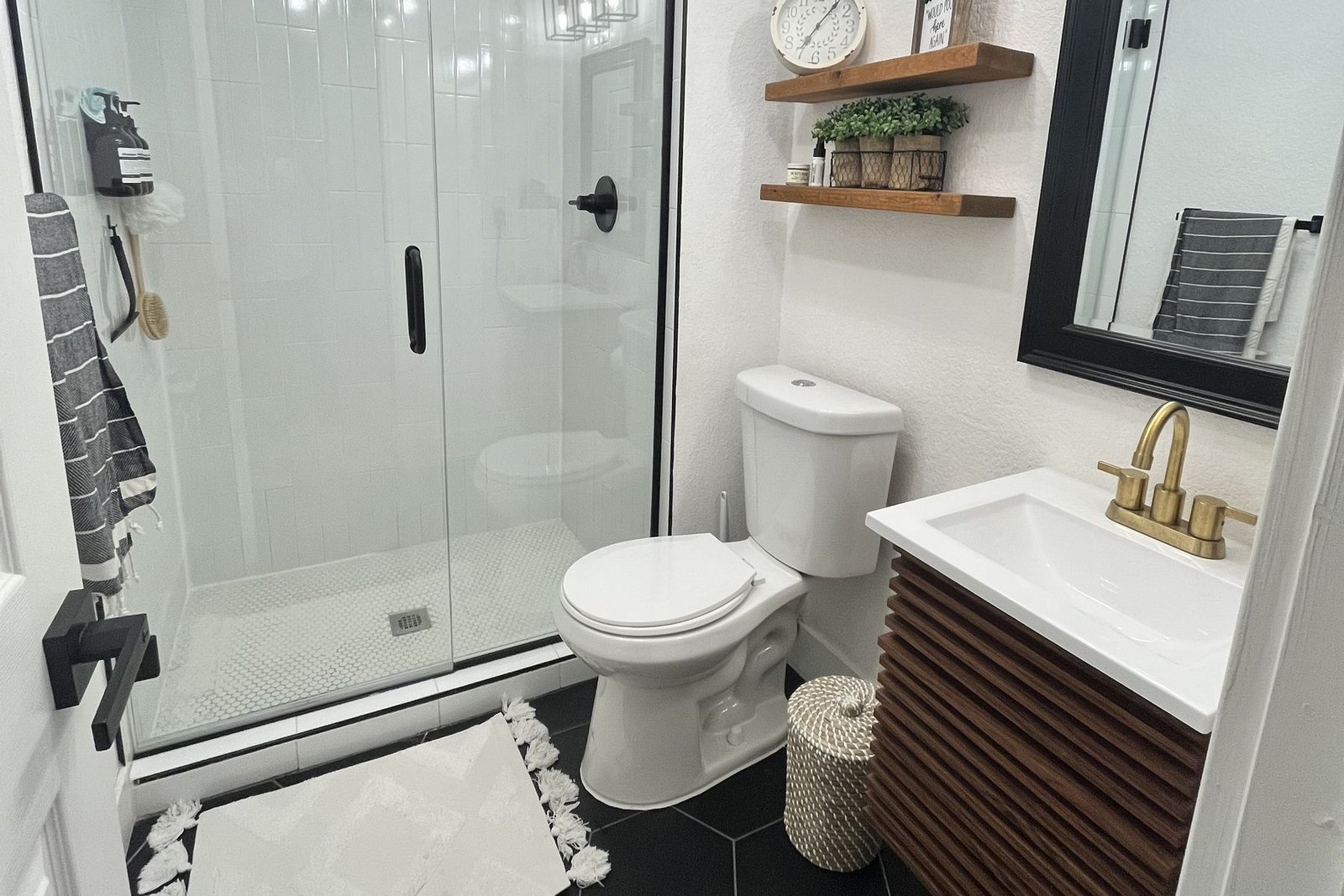 A finished small bathroom with vertically stacked white tile in the shower behind a glass panel, black hexagon floor tile, a fluted wood vanity with a brass faucet and a black framed mirror