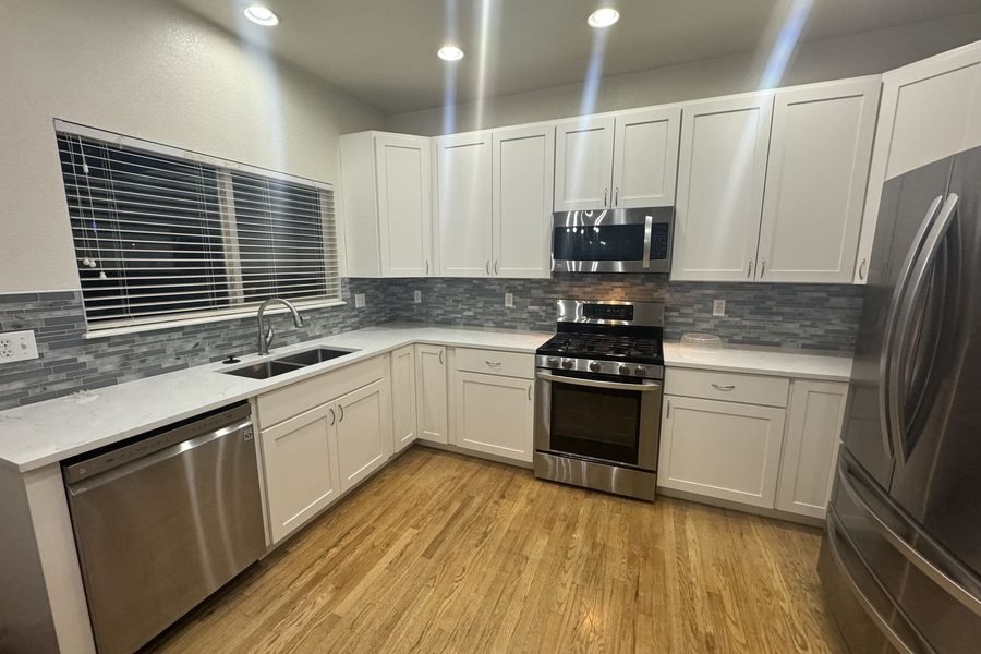 A finished kitchen with white shaker cabinets, light quartz counters, a grey linear mosaic tile backsplash, a stainless range, microwave and dishwasher, and a light oak floor