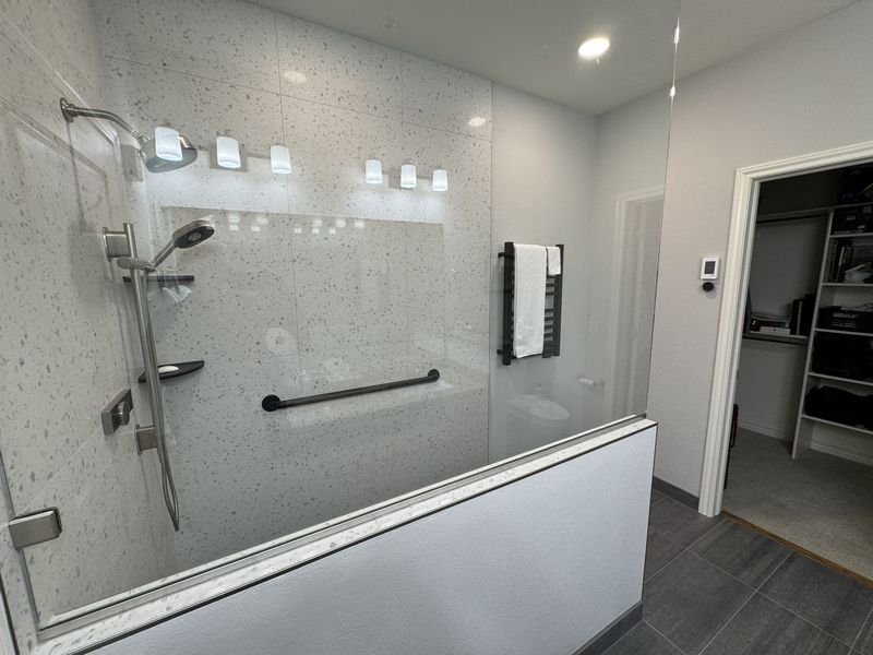 The same bathroom after: one long walk-in shower in speckled large-format tile with a full-width niche, a fixed head and a handheld on a slide bar, a black grab bar, and frameless glass sitting on a low half wall