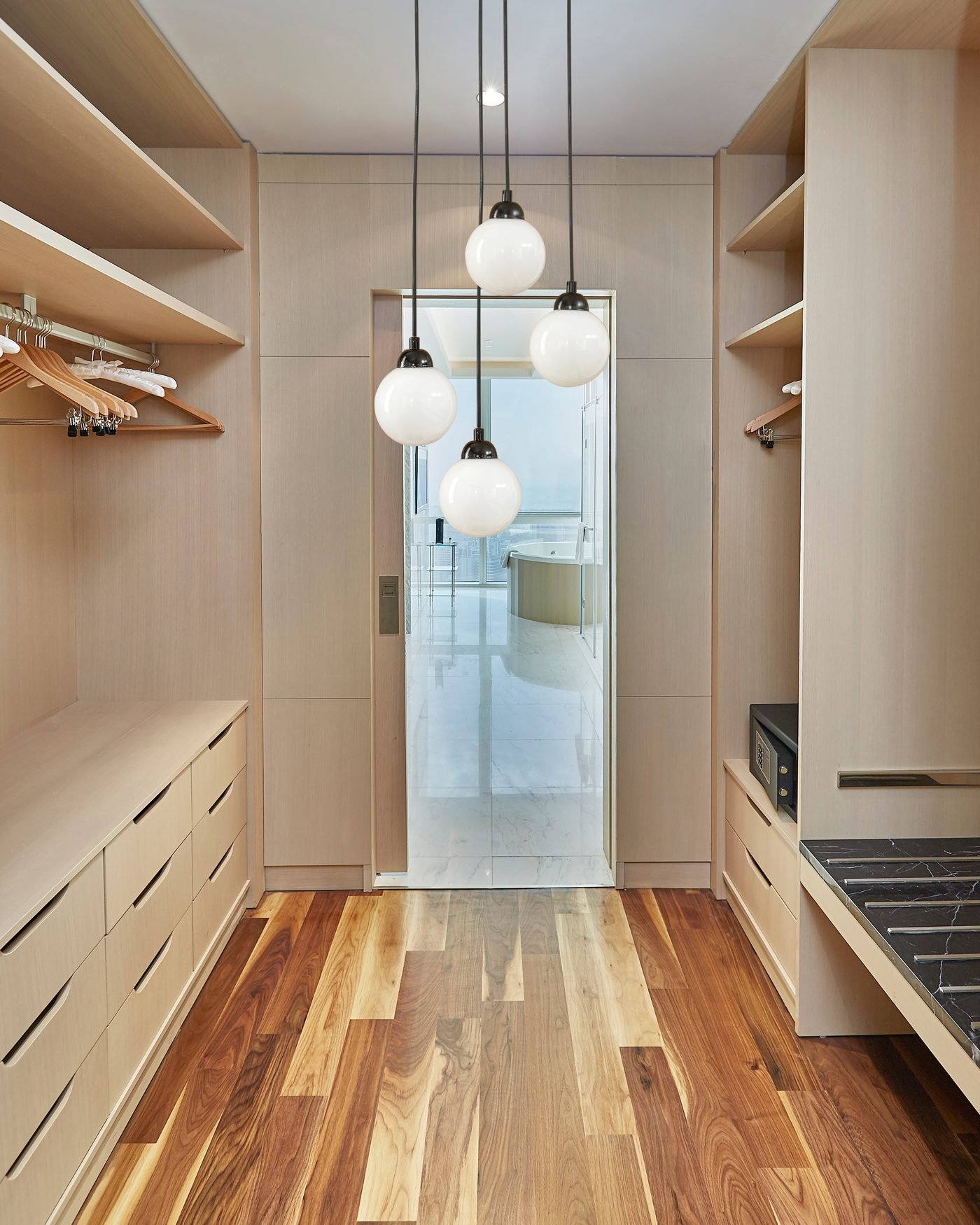 A galley walk in closet in pale oak, with drawer banks, open shelving and hanging rails down both sides under four globe pendants, over a walnut plank floor
